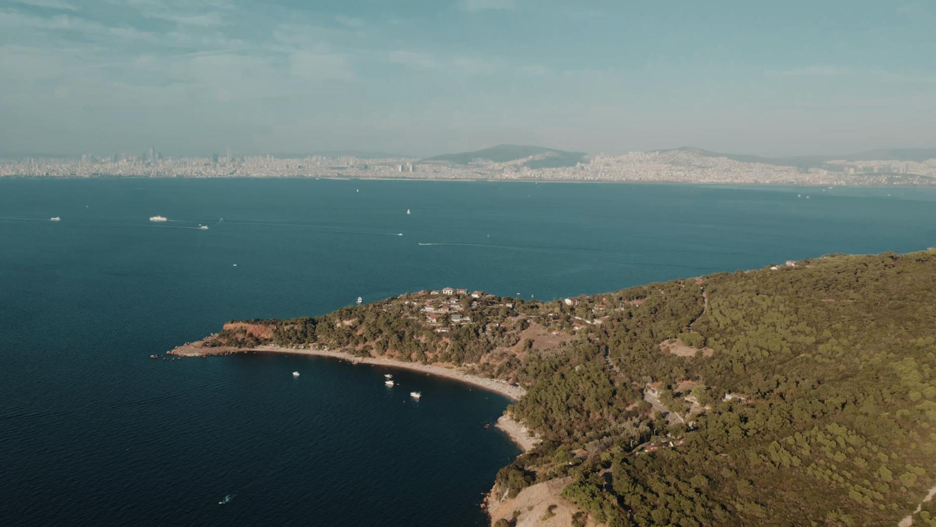 Vue aérienne de la partie boisée de l'île de Burgazada, avec une petite crique et des maisons, le tout surplombant la mer de Marmara avec la ligne d'horizon d'Istanbul en arrière-plan.