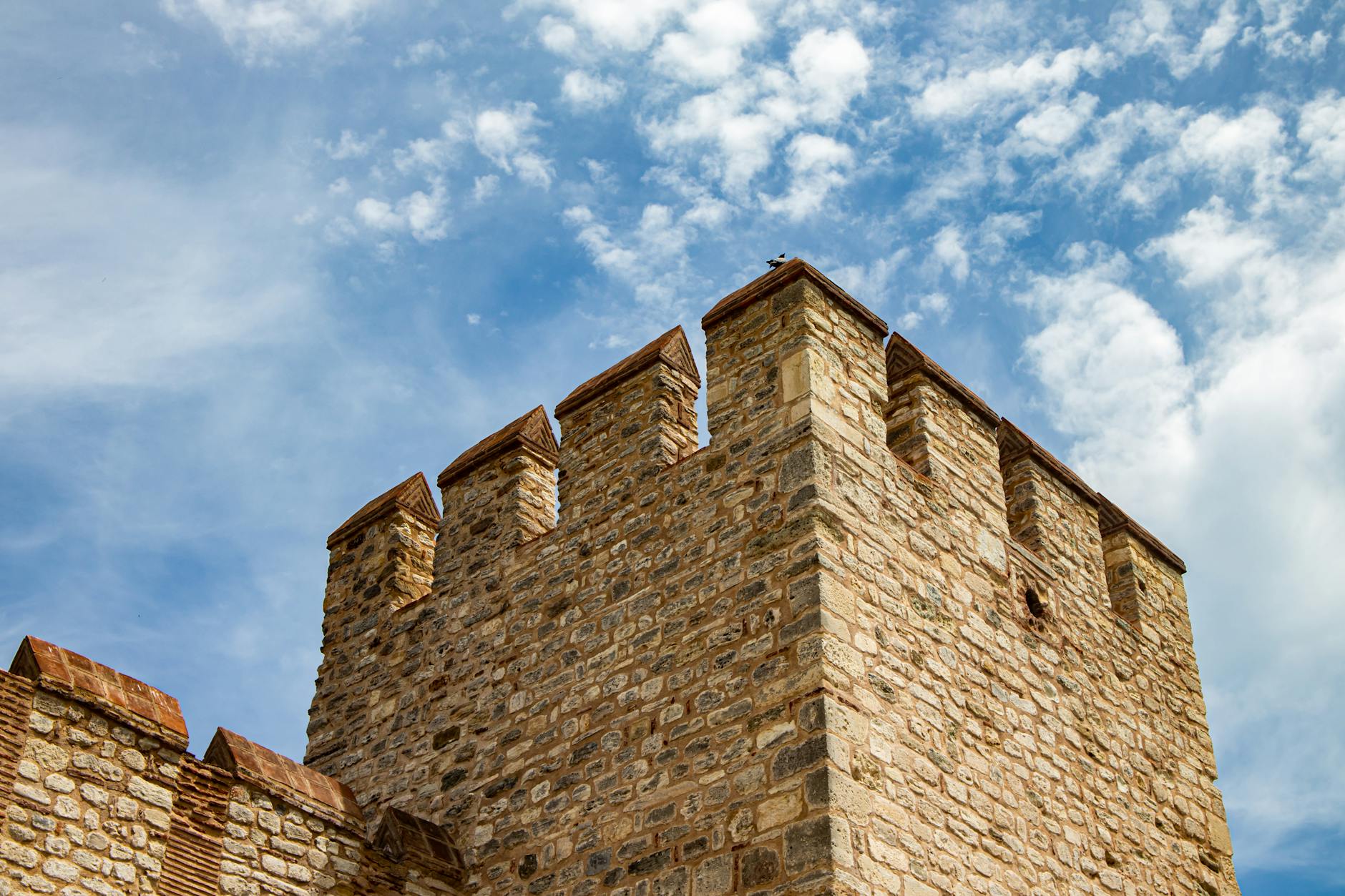 Gros plan en contre-plongée sur les créneaux et la maçonnerie en pierre des anciennes murailles de Théodose à Istanbul, sous un ciel bleu vif parsemé de nuages blancs. Cette vue évoque une longue marche solitaire le long de ces fortifications byzantines historiques.