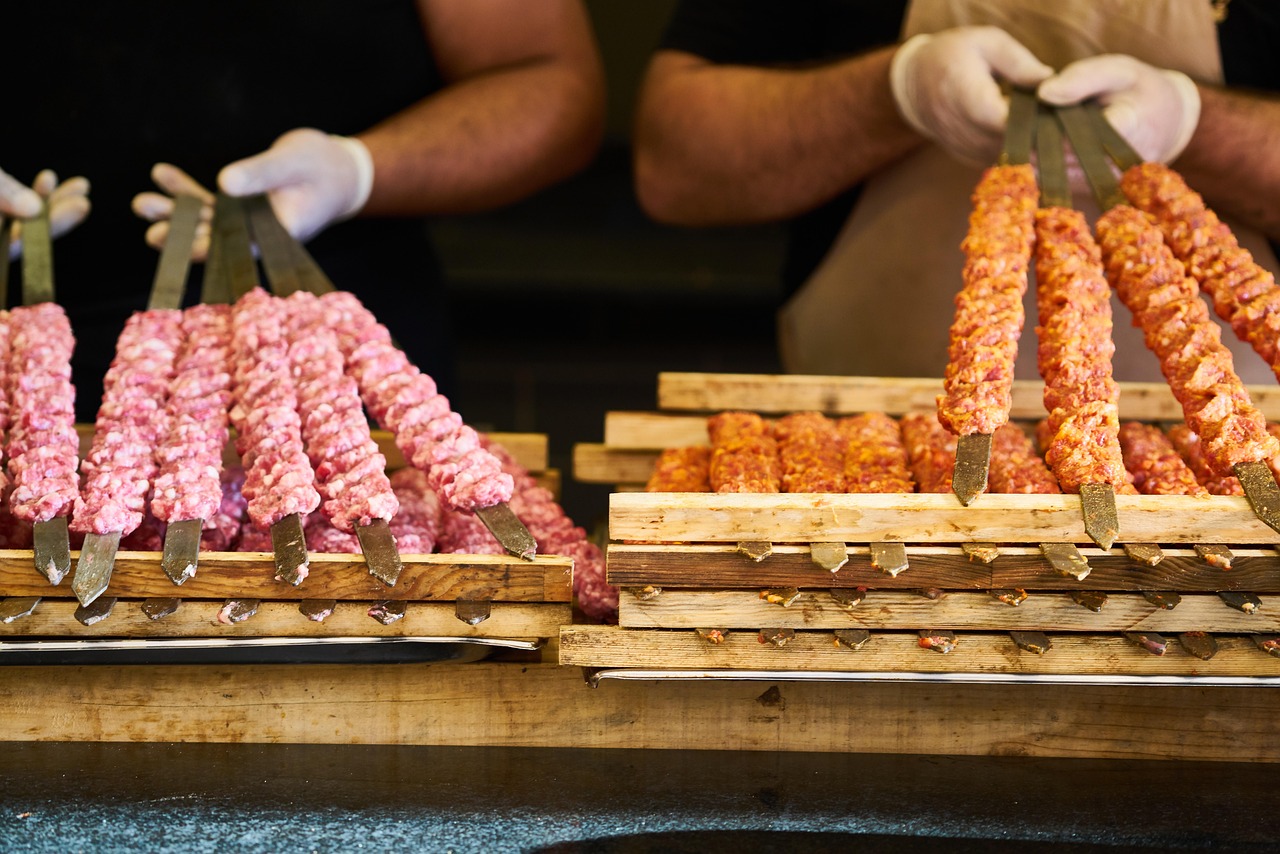 Gros plan sur des brochettes de viande hachée crue, certaines de couleur rose et d'autres épicées en rouge-orange, prêtes à griller. Des mains gantées de blanc préparent ou arrangent ces brochettes sur des supports en bois, illustrant l’Art de l’Ocakbaşı et le rituel avant la grillade au feu de bois, typique de la cuisine turque.
