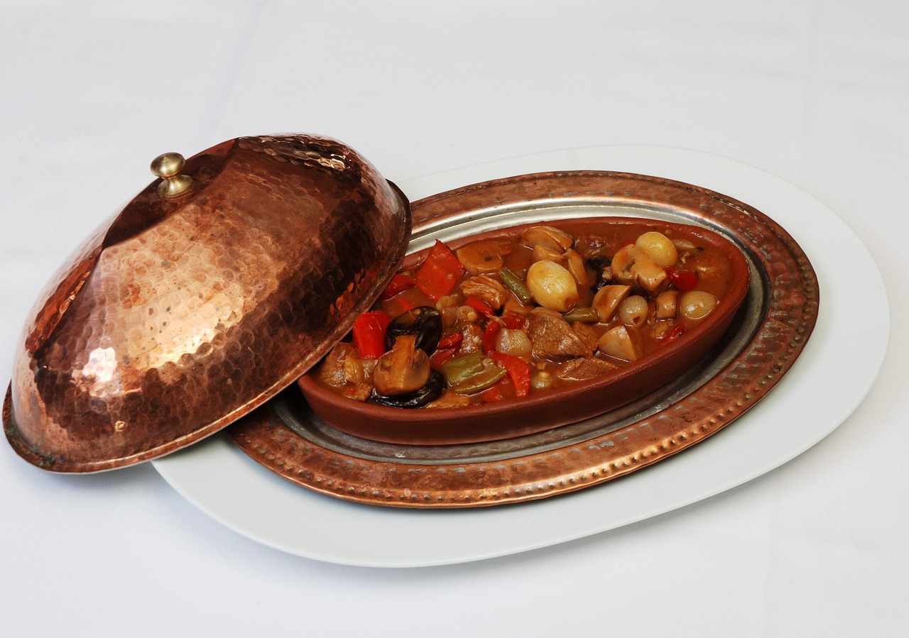 Gros plan sur un plat de ragoût traditionnel turc servi dans un récipient en terre cuite (güveç) rempli de morceaux de viande, champignons et légumes, présenté sur un plateau en cuivre martelé typique de la cuisine de l'Ocakbaşı. Le couvercle en cuivre brillant repose à côté, évoquant l'atmosphère rustique et authentique du rituel de la grillade au feu de bois.